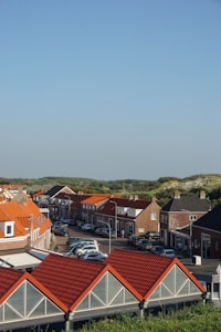 A picturesque residential neighborhood features rows of houses with distinct red-tiled roofs and brick exteriors. The street is lined with parked cars, and lush green hills rise in the background under a clear blue sky.