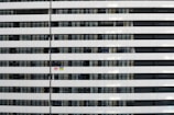 A modern multi-story building facade featuring a repetitive pattern of horizontal white and dark sections. Multiple windows are visible, some with curtains drawn. Two flags, one Georgian and one Ukrainian, are displayed in one of the windows.