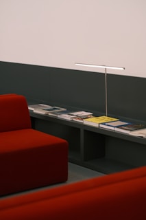 A minimalist interior setting featuring deep red, modern couches alongside a sleek shelf displaying a variety of books. A slim, long lamp provides a touch of modern elegance in the space, highlighting the books beneath.