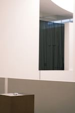 An interior space with minimalistic design, featuring smooth, pale-colored walls and a dark curtain in the background. A small sculpture or object is placed on a pedestal in the foreground, surrounded by clean lines and a modern aesthetic.
