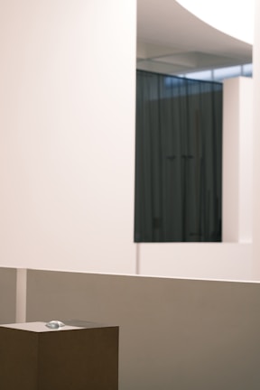 An interior space with minimalistic design, featuring smooth, pale-colored walls and a dark curtain in the background. A small sculpture or object is placed on a pedestal in the foreground, surrounded by clean lines and a modern aesthetic.