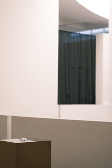 An interior space with minimalistic design, featuring smooth, pale-colored walls and a dark curtain in the background. A small sculpture or object is placed on a pedestal in the foreground, surrounded by clean lines and a modern aesthetic.