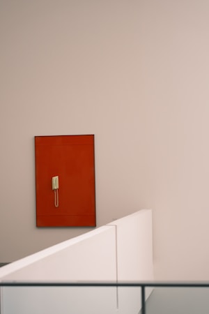 A minimalist indoor setting with a beige wall. A vintage telephone is mounted on a contrasting red rectangular board affixed to the wall. The foreground features clean, white architectural lines forming a barrier, contributing to the modern aesthetic.