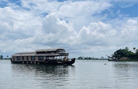 A traditional houseboat floats on calm waters under a partly cloudy sky. There are lush green trees on the shoreline in the distance, creating a tranquil and scenic view. The boat has a thatched roof and multiple windows, adding to its rustic charm.
