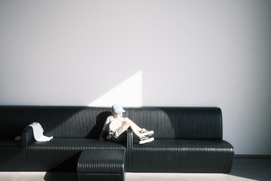 A person is sitting on a modern, ribbed black sofa positioned against a smooth, white wall. The person appears relaxed and is bathed in a patch of sunlight creating a stark contrast against the shadowy part of the sofa. There is a piece of white clothing or fabric draped over the armrest beside them.