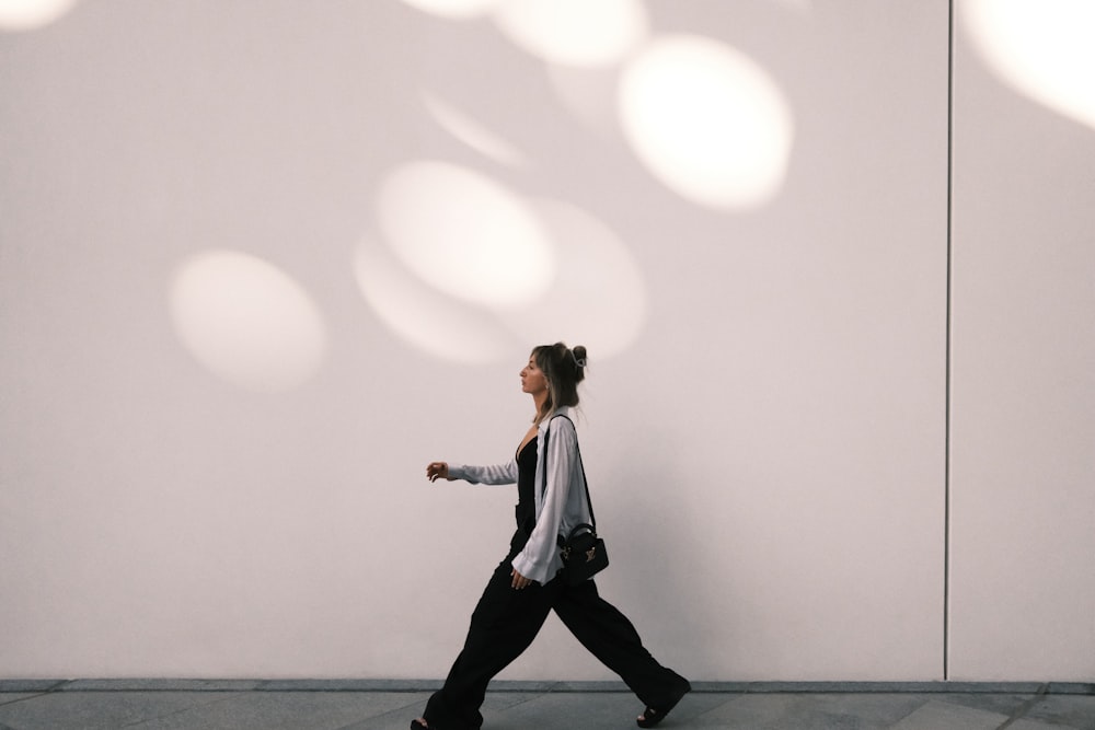 a woman walking down a sidewalk past a white wall