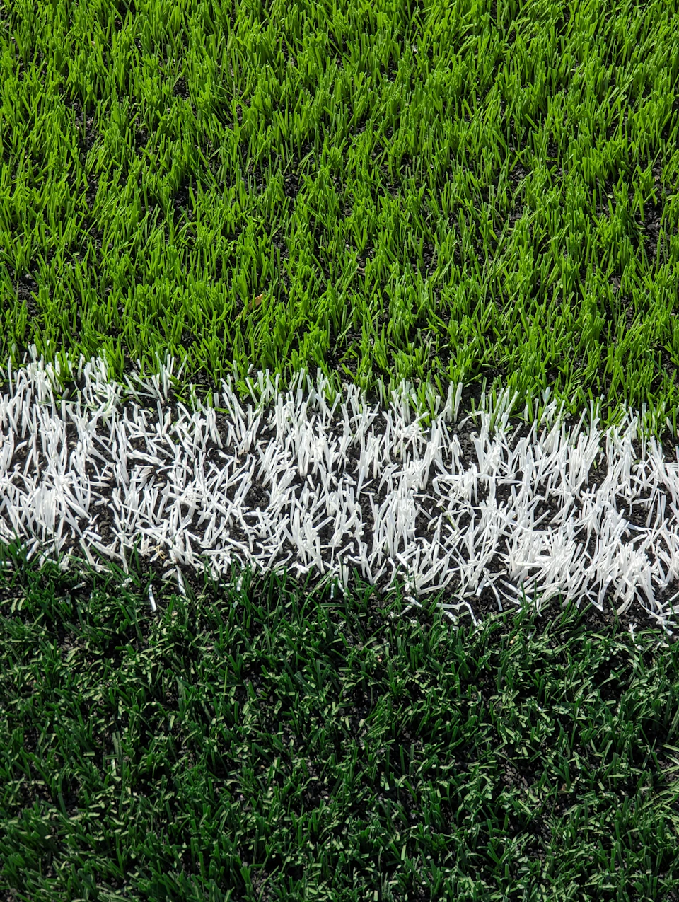 The image shows a close-up view of a soccer field, with its green grass and white lines marking the boundaries. The field is divided into three distinct sections by two parallel white lines running horizontally across it. The top section has a darker shade of green compared to the bottom section, creating a sense of depth in the image. The perspective suggests that the photo was taken from the side or slightly above the field, providing an overview of its layout and structure.