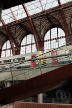 A modern train station interior with large iron and glass archways. A train with red and white color scheme is seen on the elevated tracks, with a sloped walkway in the foreground. The architecture is spacious and industrial, featuring extensive use of glass and metal.