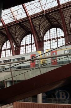 A modern train station interior with large iron and glass archways. A train with red and white color scheme is seen on the elevated tracks, with a sloped walkway in the foreground. The architecture is spacious and industrial, featuring extensive use of glass and metal.