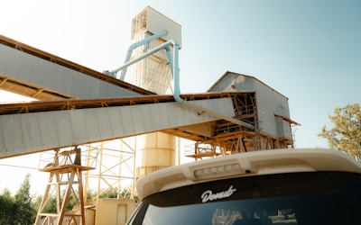 An industrial setting with a large metal structure that includes conveyor belts and piping, set against a clear sky. The foreground features the rear of a vehicle with a sticker on the window.