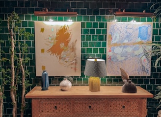 A wooden console table is adorned with a decorative lamp, two vases, and a sculptural object. Above the table hang two abstract paintings, illuminated by overhead lights. The background features a dark green tile wall with some greenery on the sides.