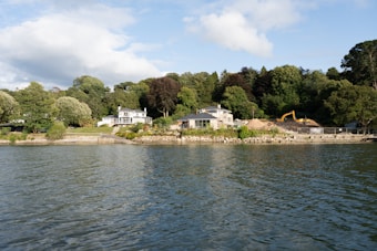 A scenic waterfront landscape featuring a couple of houses surrounded by lush green trees. The houses are situated near a shoreline with clear, calm water in the foreground. A yellow excavator is visible at a construction site next to one of the houses, indicating some development work. The sky is partly cloudy with sunlight illuminating parts of the scene.
