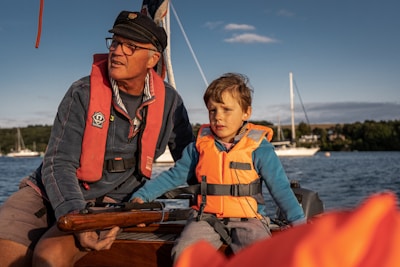 An adult and a child wearing life jackets are on a boat in a body of water. The adult is wearing glasses and a hat, holding a wooden tiller, while the child looks ahead. Sailboats are visible in the background under a clear sky.