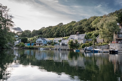 A picturesque riverside scene featuring several stone cottages and houses along the riverbank, surrounded by lush greenery and trees. The calm water reflects the buildings and the sky, adding to the serene atmosphere. There are small boats moored along the shore nearby.
