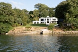 A modern white house with large windows is situated amid lush greenery. The house is surrounded by a well-maintained lawn that slopes down to a stone-lined waterfront. Dense trees encompass the property, creating a serene and secluded atmosphere.