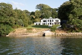 A modern white house with large windows is situated amid lush greenery. The house is surrounded by a well-maintained lawn that slopes down to a stone-lined waterfront. Dense trees encompass the property, creating a serene and secluded atmosphere.