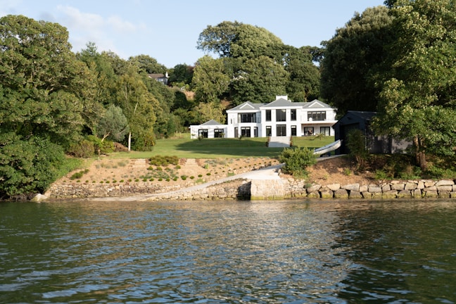 A modern white house with large windows is situated amid lush greenery. The house is surrounded by a well-maintained lawn that slopes down to a stone-lined waterfront. Dense trees encompass the property, creating a serene and secluded atmosphere.