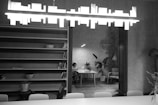 A modern, minimalist interior features a long bookshelf with a few books and objects. A stylish pendant light hangs from the ceiling. In the background, a person sits at a table with some potted plants around, creating an inviting and cozy atmosphere. The room has an industrial touch with exposed brick detailing.