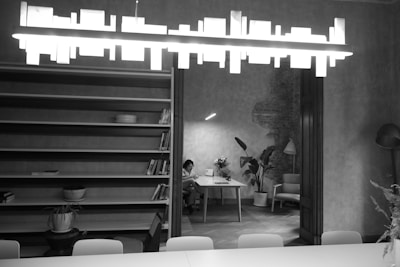 A modern, minimalist interior features a long bookshelf with a few books and objects. A stylish pendant light hangs from the ceiling. In the background, a person sits at a table with some potted plants around, creating an inviting and cozy atmosphere. The room has an industrial touch with exposed brick detailing.