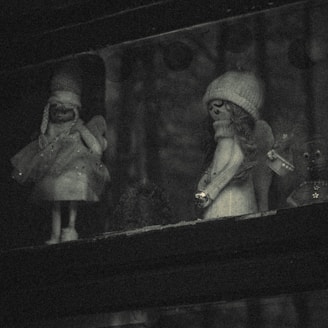 Two small figurines are placed in a dimly lit area, each resembling whimsical characters with accessories like scarves and hats. The scene appears to have a gentle, vintage aesthetic with muted lighting.