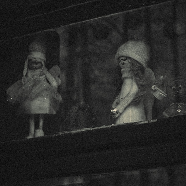 Two small figurines are placed in a dimly lit area, each resembling whimsical characters with accessories like scarves and hats. The scene appears to have a gentle, vintage aesthetic with muted lighting.