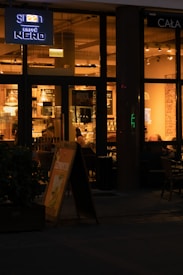 A warmly lit café interior seen through large windows, with a few people sitting inside. The café is named Caffè Nero, indicated by a neon sign. A sandwich board displaying a menu item stands outside near a plant in a pot. The interior features hanging lights and a cozy atmosphere.