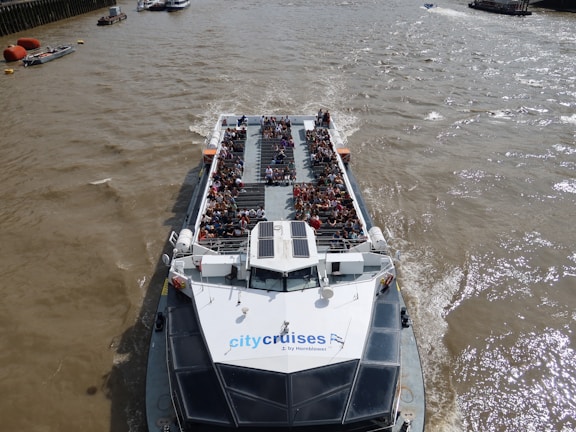 A river cruise boat is filled with passengers enjoying a scenic trip. The boat is on a wide river with a few smaller boats visible in the distance. The deck of the cruise boat is crowded with people sitting and observing the surroundings. The water is a murky brown, indicating the river's active currents.