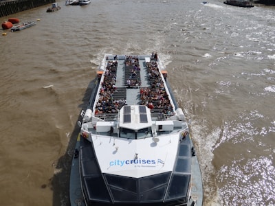 A river cruise boat is filled with passengers enjoying a scenic trip. The boat is on a wide river with a few smaller boats visible in the distance. The deck of the cruise boat is crowded with people sitting and observing the surroundings. The water is a murky brown, indicating the river's active currents.