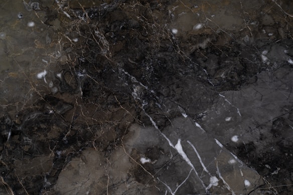 A dark marble surface featuring intricate patterns and veining with a combination of earthy and white tones. The texture is smooth with natural cracks and a polished finish.