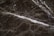 A close-up view of a dark marble surface featuring intricate white veining patterns. The texture appears smooth with a glossy finish. The marble displays a natural, earthy color palette.