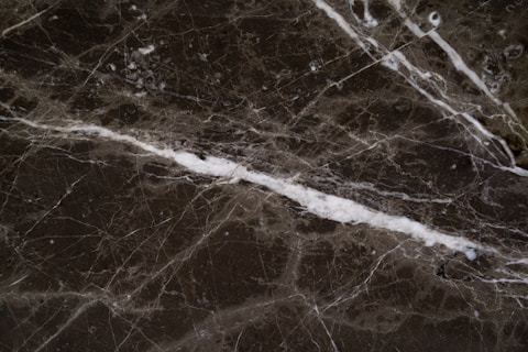 A close-up view of a dark marble surface featuring intricate white veining patterns. The texture appears smooth with a glossy finish. The marble displays a natural, earthy color palette.