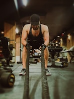 A man wearing athletic gear, including a black tank top, cap, and gloves, is engaged in an intense workout using battle ropes in a gym setting. The background shows various gym equipment such as dumbbells and machines, with a warm, low-lit ambiance.