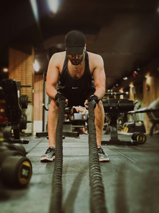 A man wearing athletic gear, including a black tank top, cap, and gloves, is engaged in an intense workout using battle ropes in a gym setting. The background shows various gym equipment such as dumbbells and machines, with a warm, low-lit ambiance.