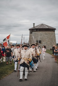 A group of men dressed in historical military uniforms is marching along a road. They are carrying flags and weapons, with onlookers present in the background. A stone fortification is visible nearby under a cloudy sky.