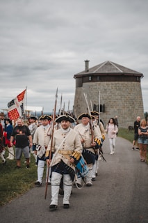 A group of men dressed in historical military uniforms is marching along a road. They are carrying flags and weapons, with onlookers present in the background. A stone fortification is visible nearby under a cloudy sky.
