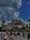 A large crowd of people walks towards a historic building with a large domed roof and minarets, set against a backdrop of partly cloudy skies. The scene is lively with tourists and locals enjoying the area, surrounded by lush greenery and palm trees.