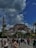 A large crowd of people walks towards a historic building with a large domed roof and minarets, set against a backdrop of partly cloudy skies. The scene is lively with tourists and locals enjoying the area, surrounded by lush greenery and palm trees.