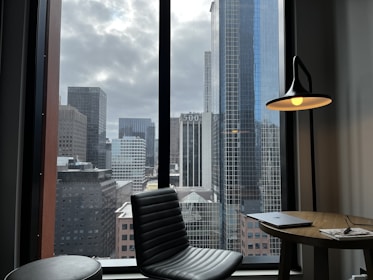A modern office or living space with a view of a cityscape filled with tall, glass skyscrapers. The room features a black leather chair and a wooden table with a closed laptop and papers. A stylish floor lamp with a glowing bulb adds warmth to the industrial setting.