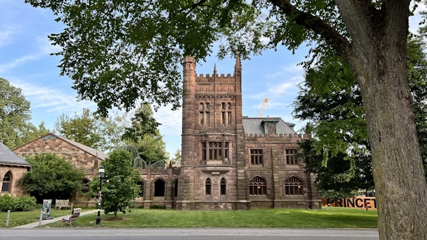 A historic building made of stone with a tall tower and arched windows, situated in a lush green area with trees and a walkway in the foreground. The structure has an old architectural style, possibly a university or institutional building.