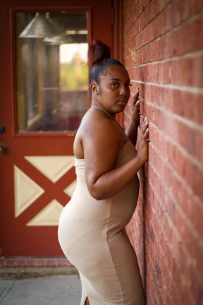 A young woman wearing a sleek, beige dress standing against a clean white wall.