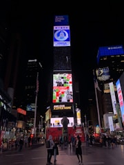 A busy urban scene featuring the iconic Times Square in New York City. Bright electronic billboards with advertisements, including prominent brands such as Coca-Cola, illuminate the area. People walk around and gather on the red steps beneath the billboards, creating a dynamic and lively atmosphere.
