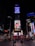 A busy urban scene featuring the iconic Times Square in New York City. Bright electronic billboards with advertisements, including prominent brands such as Coca-Cola, illuminate the area. People walk around and gather on the red steps beneath the billboards, creating a dynamic and lively atmosphere.