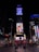 A busy urban scene featuring the iconic Times Square in New York City. Bright electronic billboards with advertisements, including prominent brands such as Coca-Cola, illuminate the area. People walk around and gather on the red steps beneath the billboards, creating a dynamic and lively atmosphere.