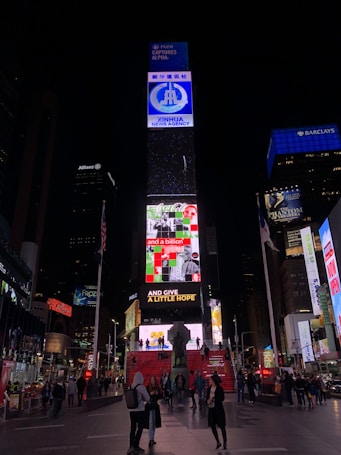 A busy urban scene featuring the iconic Times Square in New York City. Bright electronic billboards with advertisements, including prominent brands such as Coca-Cola, illuminate the area. People walk around and gather on the red steps beneath the billboards, creating a dynamic and lively atmosphere.
