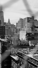 A black and white cityscape featuring urban buildings covered with graffiti. The focus is on the rooftop graffiti art with various styles and tags. In the background, several skyscrapers and a prominent tall building with a spire are visible, representing an iconic skyline.