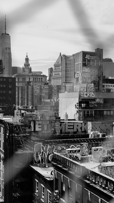 A black and white cityscape featuring urban buildings covered with graffiti. The focus is on the rooftop graffiti art with various styles and tags. In the background, several skyscrapers and a prominent tall building with a spire are visible, representing an iconic skyline.