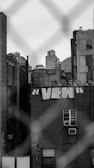 Urban scene featuring a brick building with white graffiti letters spelling 'VIEW' on its side. The image is framed by a blurred chain-link fence in the foreground. Rooftops and satellite dishes are visible on adjacent buildings, along with additional graffiti. The atmosphere appears gritty and industrial.