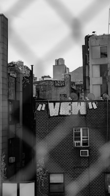 Urban scene featuring a brick building with white graffiti letters spelling 'VIEW' on its side. The image is framed by a blurred chain-link fence in the foreground. Rooftops and satellite dishes are visible on adjacent buildings, along with additional graffiti. The atmosphere appears gritty and industrial.