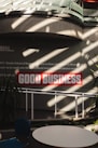 Sunlight streams through overhead beams, casting shadows on a wall featuring a prominent sign with bold red letters stating 'GOOD BUSINESS'. The environment appears to be indoors with modern architectural elements and minimalistic decor. Potted plants and furniture create a relaxed, professional atmosphere.