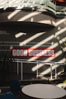 Sunlight streams through overhead beams, casting shadows on a wall featuring a prominent sign with bold red letters stating 'GOOD BUSINESS'. The environment appears to be indoors with modern architectural elements and minimalistic decor. Potted plants and furniture create a relaxed, professional atmosphere.